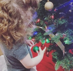 Curly brown haired two year old girl placing a green Christmas ornament on the Christmas tree.