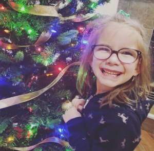 Brown haired 6 year old girl in a pony tail and glasses smiling in front of the Christmas tree.