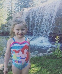 Hailey at 3 years old. She has brown hair and is wearing an Ariel themed bathing suit. She is smiling standing in front of a waterfall.
