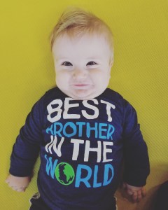 Timothy a blonde haired blue eyed 8 month old. He is smiling. His blue shirt says best brother in the world.