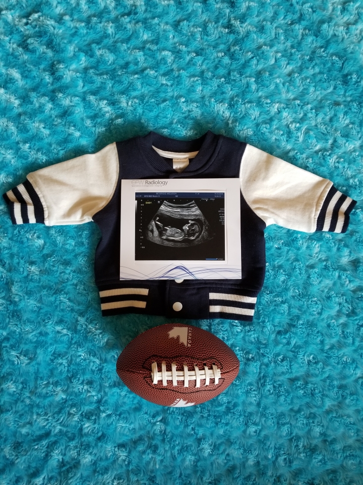 Photo of our gender announcement for Timothy. A baby blue background, a newborn size varsity jacket with an ultrasound picture on top with a baby sized football laying underneath the jacket. 