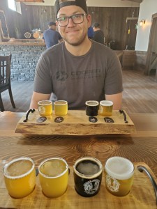 Photo of James smiling sitting at a table with two sample flights of beer in front of him from our trip.